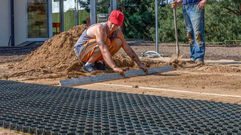 A base layer of plastic honeycomb grids is laid in the garden. The honeycomb grids lie on a bed of sand. A man is in the process of preparing the bed for the next ground grids and draws off the sand using a gauge made of pipes.