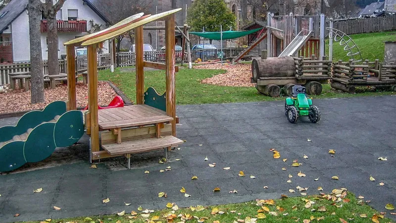 View of the central area of a playground in Olsberg with a varied and natural design. The center of the playground consists of a spacious play area made of green fall protection slabs with a small playhouse.