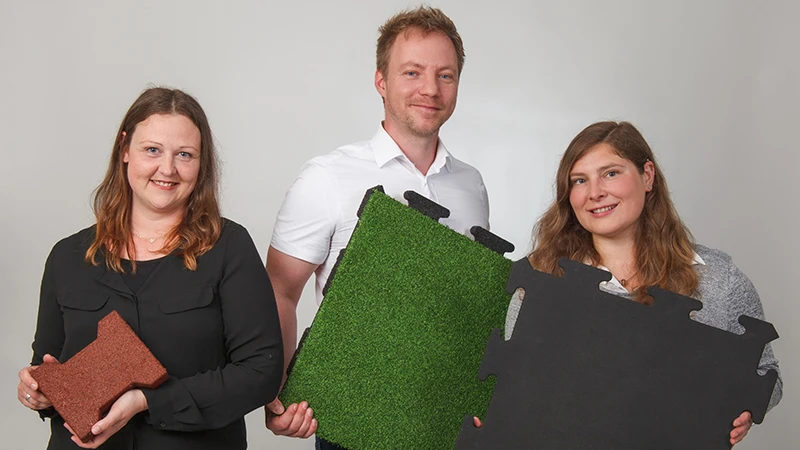 With friendly smiles, three WARCO consultants display various floor tiles made from PU-bonded rubber granulate in brown, green, and black. Ideal for balconies, these tiles provide slip-resistant, durable properties. Each surface features a unique design and is easy to install. Their expert guidance ensures the right flooring choice for a safe and attractive balcony. Low maintenance and comfortable underfoot, the tiles are a practical option for any balcony project.