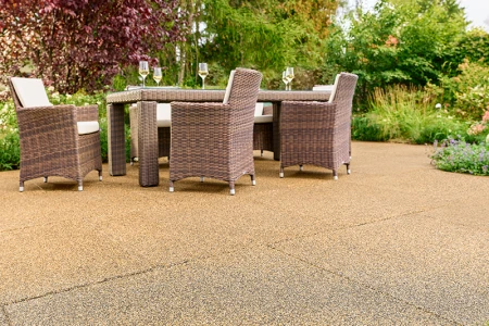 Sunlit garden patio featuring beige-brown WARCO tiles made from PU-bound rubber granules. The slip-resistant, water-permeable surface ensures a comfortable walking experience and reduces noise. A sturdy garden table with woven chairs and filled wine glasses invites relaxation. Vibrant shrubs and colorful flowers create a natural setting. This elastic flooring is frost-resistant, retains its shape, and provides long-lasting durability. Ideal for year-round use, it complements any outdoor décor.