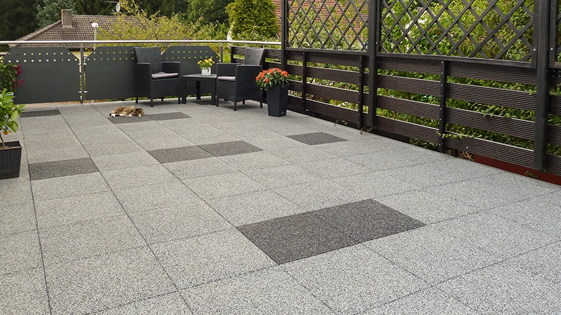 This photo shows a generously sized balcony fitted with square WARCO rubber granulate tiles in various shades of grey. A cat is resting in one corner, while comfortable seating and colourful flowers on the opposite side create an inviting ambience. The flooring is slip-resistant, weatherproof, and offers excellent sound and impact absorption. Thanks to its low-maintenance surface, it is ideal for balconies, terraces, and other outdoor areas.