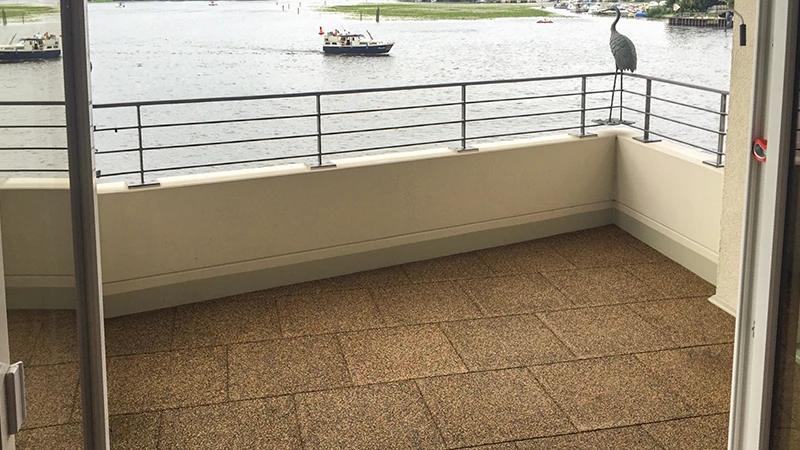 A balcony is fitted with light-brown tiles made of PU-bonded rubber granulate. The square pieces create a continuous, slip-resistant area that feels comfortable underfoot. The elastic structure reduces footstep noise and vibrations. Thanks to the open-pored surface, water drains away easily, providing safety in rainy conditions. The warm colour blends with the light railing and adds a welcoming touch. In the background, a body of water is visible with boats along its shore.