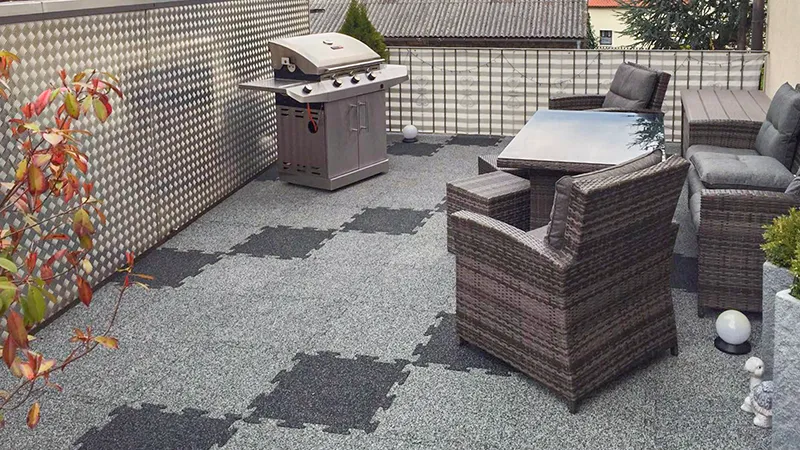 Shown here is a spacious rooftop terrace featuring grey WARCO interlocking puzzle tiles. Their slip-resistant surface ensures secure footing and comfortable walking. Complementing this setup are weatherproof rattan chairs and a barbecue grill, perfect for social gatherings. The durable flooring is easy to install and an excellent choice for outdoor spaces, thanks to its low-maintenance and weather-resistant properties. Enjoy a welcoming area for relaxation and entertaining.