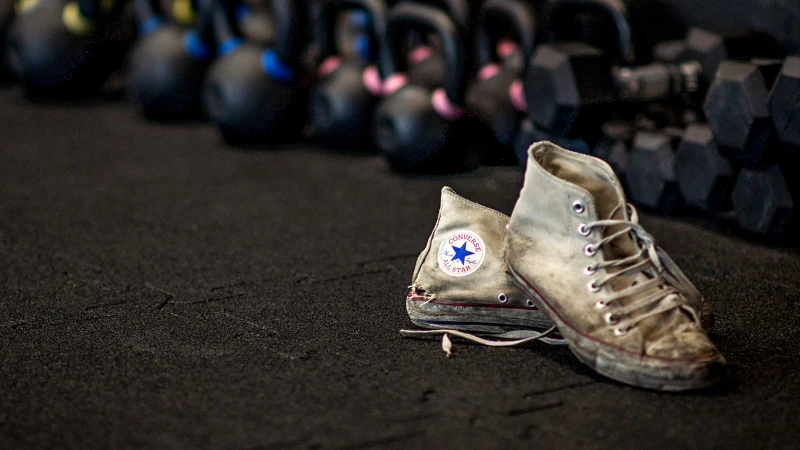 A pair of heavily used Converse ALL STAR sneakers stands in front of a set of dumbbells in the gym on black WARCO fitness mats.