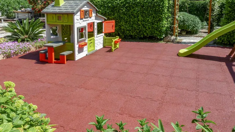 In the middle of the garden, surrounded by shrubs, there is a play area made of brick-red WARCO playground flooring. A colorful playhouse and a children's slide are available for the children to play on.