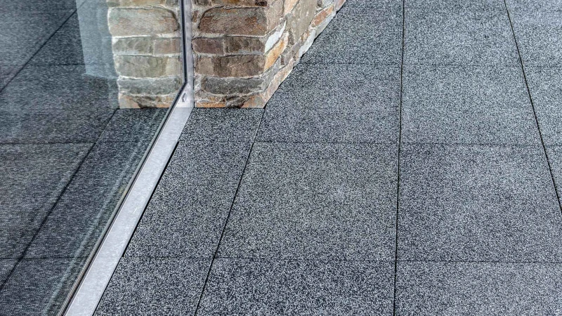 WARCO-Terrassenplatten liegen bündig an einer Hauswand aus unregelmäßig geformten Natursteinen an. Die Platten aus Gummigranulat lassen sich mit einer Stichsäge, einer Säbelsäge oder einem Messer genau und präzise zuschneiden.