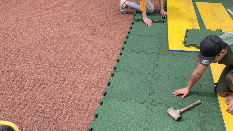A man lays green safety tiles with puzzle interlocking, i.e. a dovetail connection system formed into the sides. The safety tiles are laid on a base layer of plastic honeycomb grid. The evenly laid honeycomb grids are filled with red chippings.