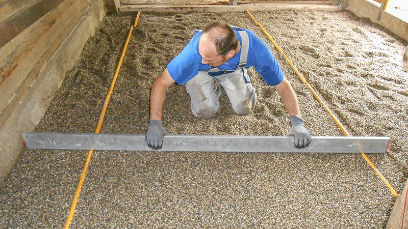 Installation of a new floor in a horse stable: A man uses a screed to level a gravel sub-base. The gravel serves as a bedding layer for the installation of a rubber granulate stable floor. The area is bordered by timber or concrete walls. The work is done with care and precision – typical for the proper preparation of a horse-friendly stable floor.