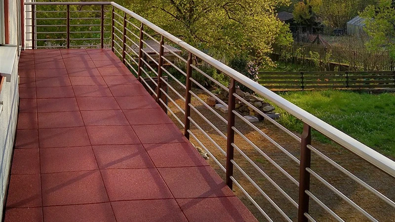 A balcony in a pleasant warm evening light is fitted with red floor tiles made of PU-bonded rubber granulate. These square rubber tiles form a slip-resistant, impact-absorbing and sound-dampening surface. Thanks to the open-pored structure, rainwater does not remain on the balcony floor. The metal railing provides an unobstructed view of the greenery.