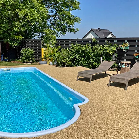 Das Bild zeigt einen Privatgarten mit einem rechteckigen Schwimmbecken. Der Boden um den Pool ist mit speziellen Schwimmbadplatten aus PU-gebundenem Gummigranulat im Format 30 x 30 x 20 mm gestaltet. Die Oberfläche der Platten besteht aus durchgefärbtem EPDM-Gummigranulat im Farbmuster Travertin, das eine natürliche Steinoptik nachempfindet. Die Platten sind fugenlos verlegt und werden durch eine fast unsichtbare Schwalbenschwanzverbindung zusammengehalten, die für eine stabile und sichere Oberfläche sorgt. Im Hintergrund steht eine gemütliche Sonnenliege, daneben grüne Büsche und ein großer Baum, der an heißen Tagen angenehmen Schatten spendet. Umgeben ist der Garten von einem modernen Holzzaun, der für Privatsphäre sorgt.