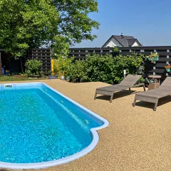 Das Bild zeigt einen Privatgarten mit einem rechteckigen Schwimmbecken. Der Boden um den Pool ist mit speziellen Schwimmbadplatten aus PU-gebundenem Gummigranulat im Format 30 x 30 x 20 mm gestaltet. Die Oberfläche der Platten besteht aus durchgefärbtem EPDM-Gummigranulat im Farbmuster Travertin, das eine natürliche Steinoptik nachempfindet. Die Platten sind fugenlos verlegt und werden durch eine fast unsichtbare Schwalbenschwanzverbindung zusammengehalten, die für eine stabile und sichere Oberfläche sorgt. Im Hintergrund steht eine gemütliche Sonnenliege, daneben grüne Büsche und ein großer Baum, der an heißen Tagen angenehmen Schatten spendet. Umgeben ist der Garten von einem modernen Holzzaun, der für Privatsphäre sorgt.