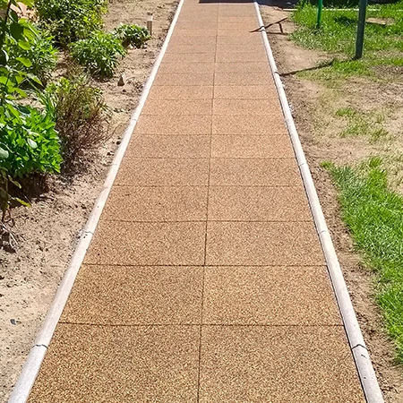 A straight garden path made of square rubber tiles with side kerb edging runs through the planting area. The brown tiles made of rubber granulate provide a low-maintenance, weather-resistant and non-slip surface. The path ensures clean, safe walking in all weathers and clearly separates the planted beds from the lawn. Ideal for gardens, courtyards and access ways.