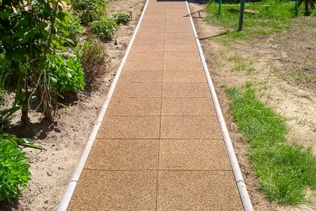 A straight garden path made of square rubber tiles with side kerb edging runs through the planting area. The brown tiles made of rubber granulate provide a low-maintenance, weather-resistant and non-slip surface. The path ensures clean, safe walking in all weathers and clearly separates the planted beds from the lawn. Ideal for gardens, courtyards and access ways.