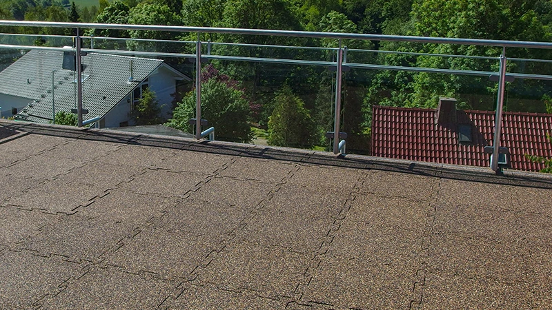 This photo features a large balcony with a clear balustrade, overlooking nearby rooftops and lush greenery. The floor is covered with WARCO puzzle tiles made from rubber granulate in a warm brown shade. Their interlocking edges create a seamless surface that is both slip-resistant and weatherproof—ideal for outdoor spaces. In addition, the flooring offers shock absorption and effective sound insulation. Thanks to its low-maintenance surface, it is a durable and comfortable choice for balconies and terraces.